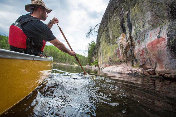 Man in a canoe paddling along a rock face with a pictograph on it