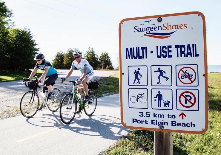 Two cyclists pedalling by Saugeen Shores Multi-Use Trail sign.