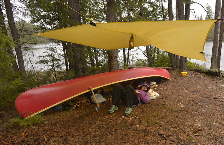 Person napping under a red canoe with a dog as a pillow