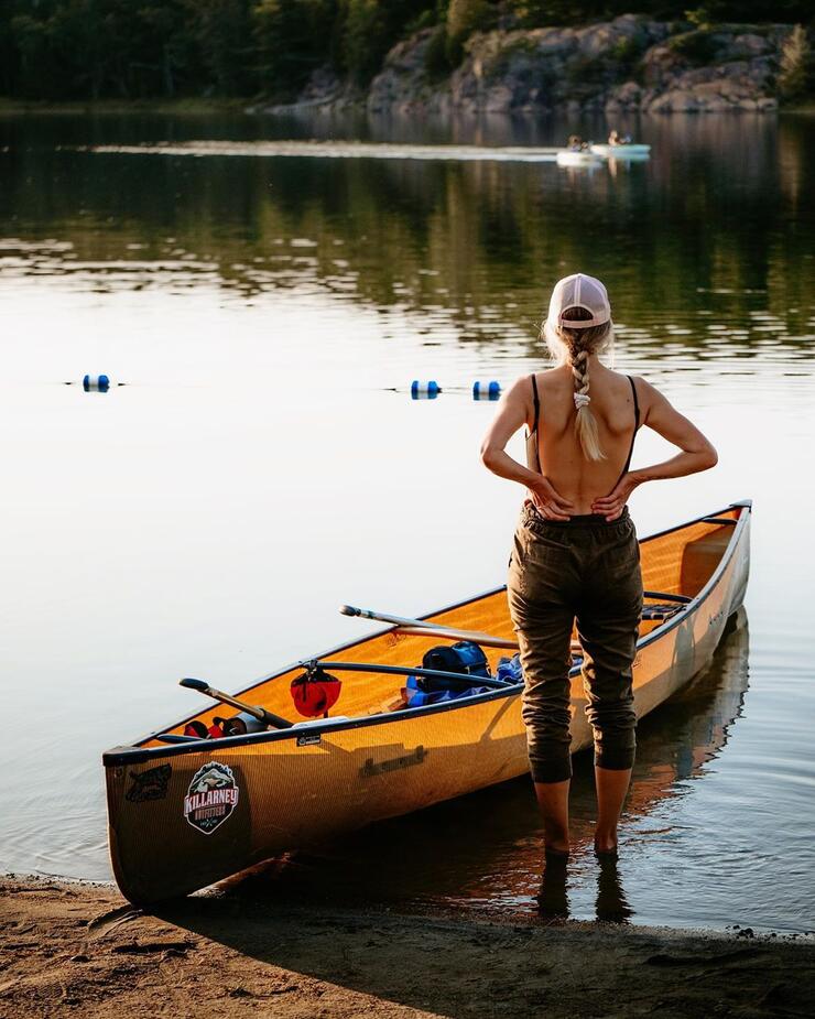 Slender, fit woman standing beside a canoe on a beach. 