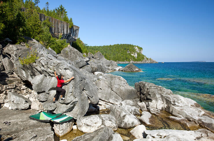 Woman climbing a boulder beside turquoise water.