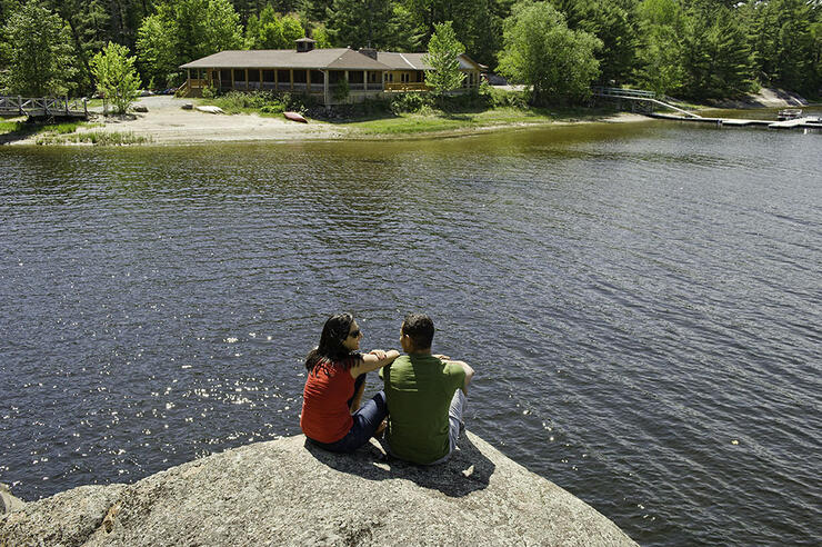 Man and woman sit on a rock by the water across from a large cottage or lodge building