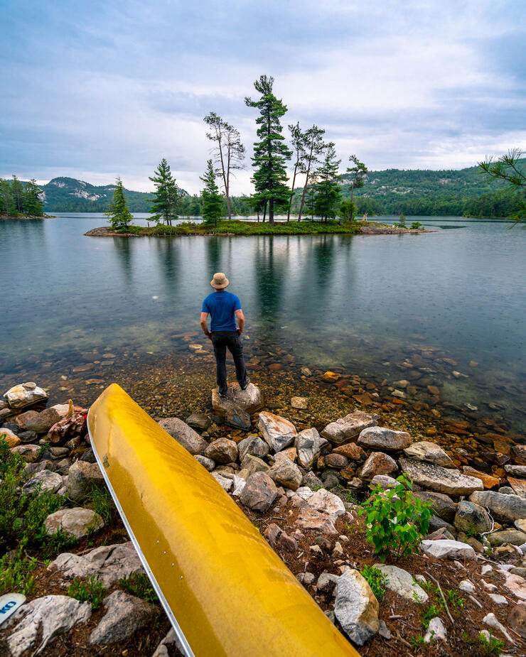 Man in straw hat stands in front of canoe at water's edge in front of small Killarney island