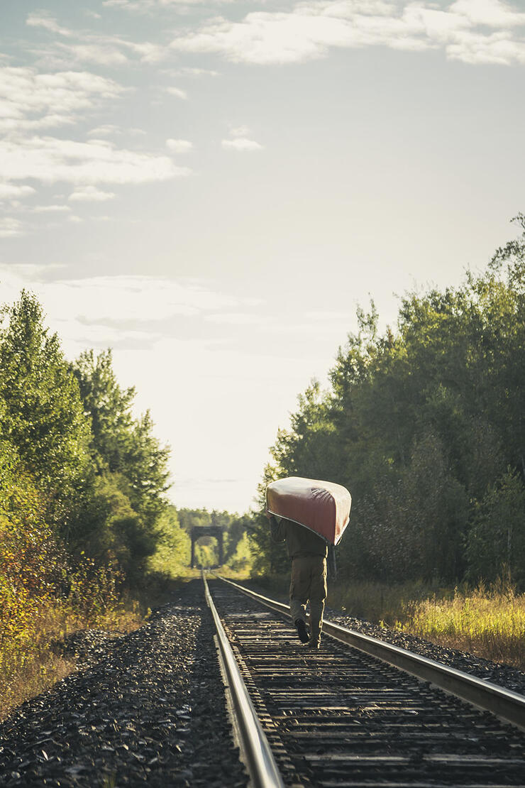 Man walking on train tracks carrying a canoe over this head
