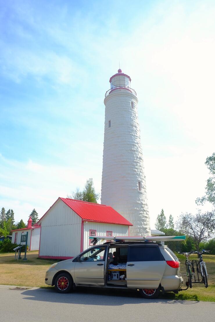 Minivan with paddleboard on roof in front of lighthouse.
