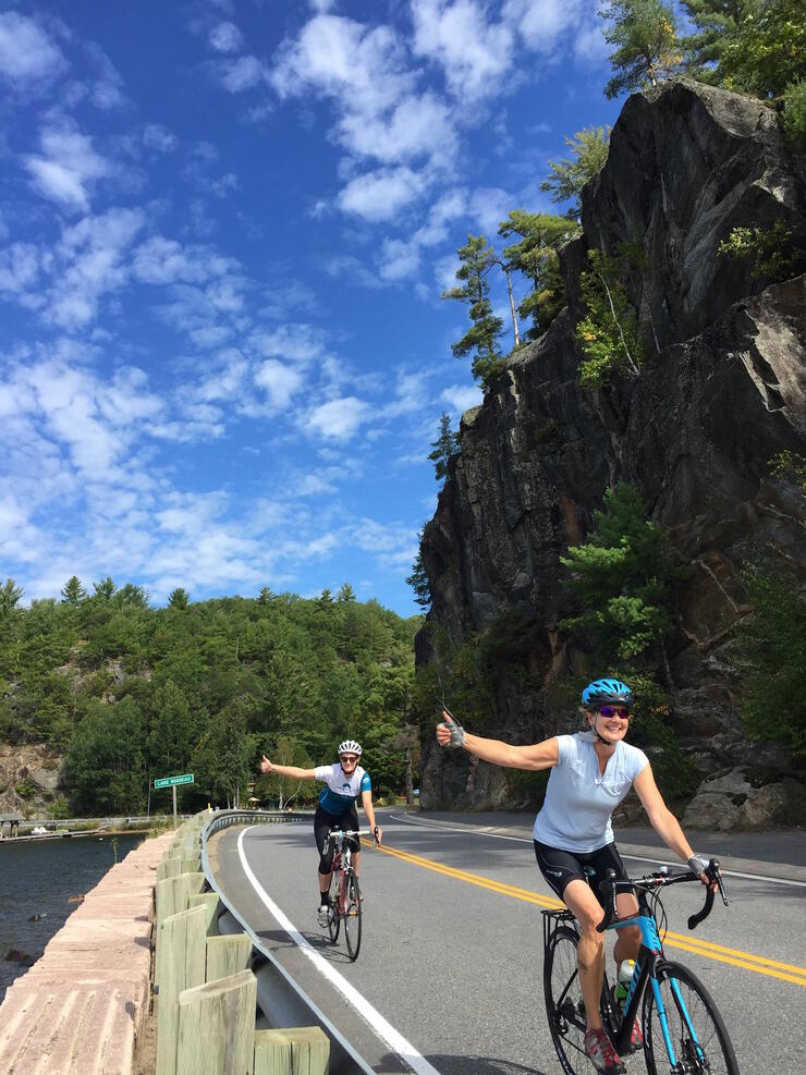 Two cyclists riding on paved road beside a lake. 