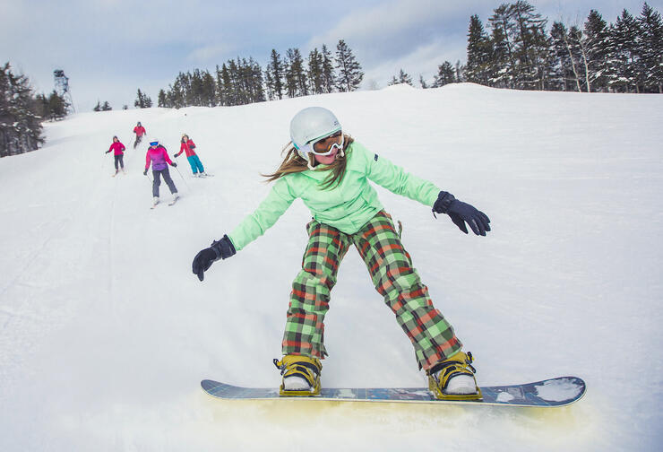Girl on snowboard with 4 skiers on slope in background