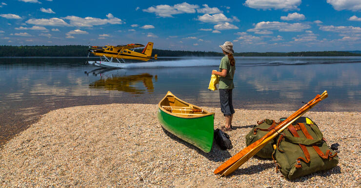 Woman standing beside green canoe on a beach in front of a yellow floatplane landing on lake