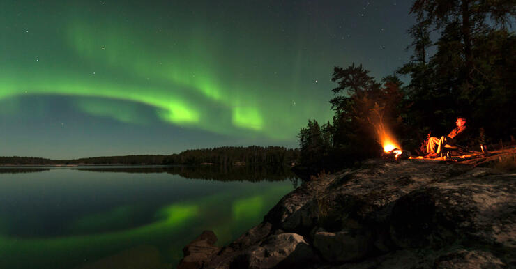 Man sitting by campfire on shore of lake looking at Aurora Borealis