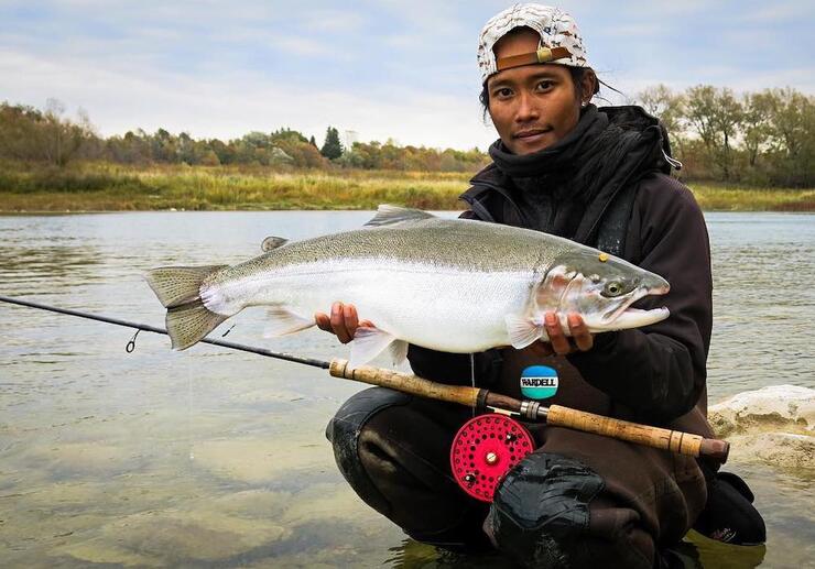 Man crouched on riverbank holding fish.