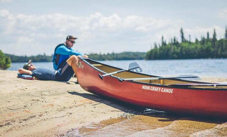 Two people sitting beside canoe pulled up on shore.